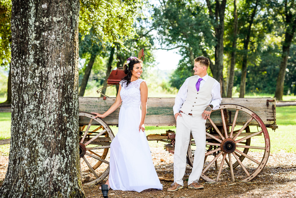 Michelle and Brent standing apart smiling at each other in front of the old wagon, Ates Ranch Wedding Barn, Rustic Barn Wedding, Pensacola wedding photographer, Lazzat Photography