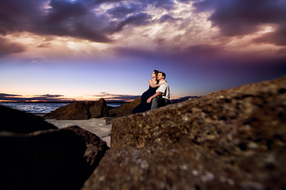 Couple sitting on rocks at the beach at sunset, Fun Engagement Session at Eden Garden and Fort Pickens, Lazzat Photography, engagement photos